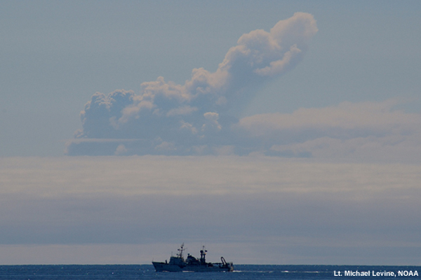 The plume of Okmock's 2008 eruption rises high above a deck of clouds over a NOAA ship sailing by.