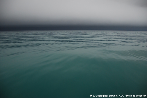 Ash is falling over the turquoise ocean waters.  The ash has formed a grey/blue cloud covering the horizon.